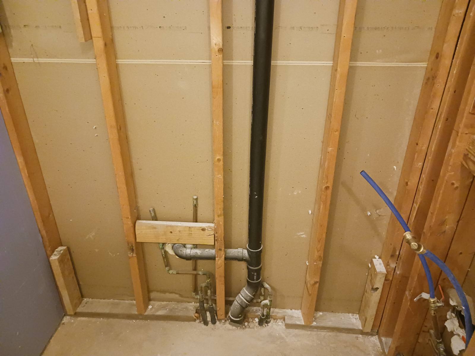 Interior wall opened up to the studs with a drain pipe running down it and blue supply lines with brass shutoff valves at the base