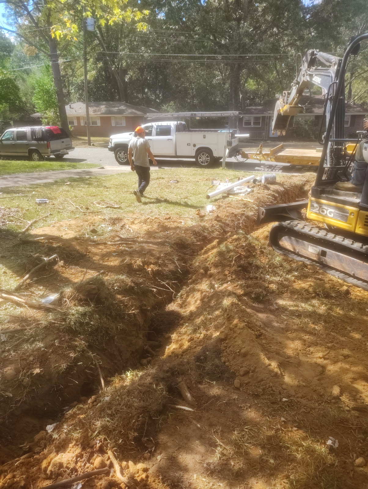 Residential yard with a freshly dug trench across the foreground, a compact excavator beside it and a person walking toward a work truck