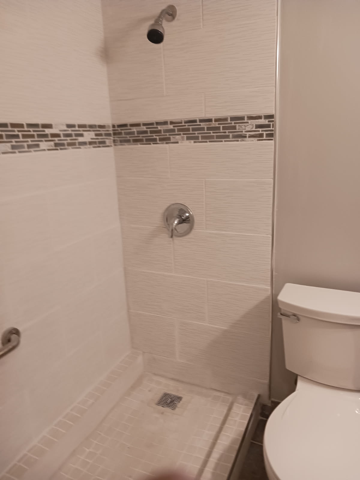 Walk-in shower finished in white textured wall tile with a mosaic accent strip, a chrome shower head and a square floor drain