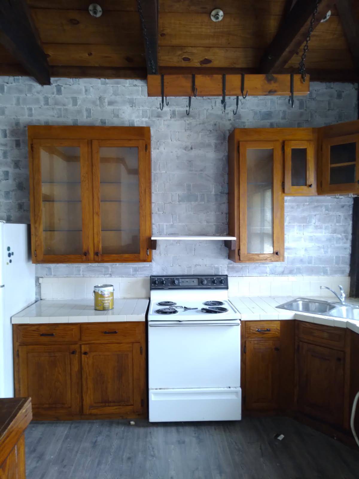 Kitchen with wooden cabinets with glass upper doors, a white stove and sink, tile counters against a brick backsplash and exposed ceiling beams