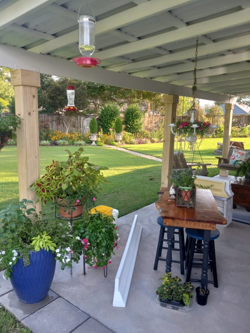 Covered patio with a light pergola ceiling over a wooden high-top table and stools, with potted plants and hanging feeders