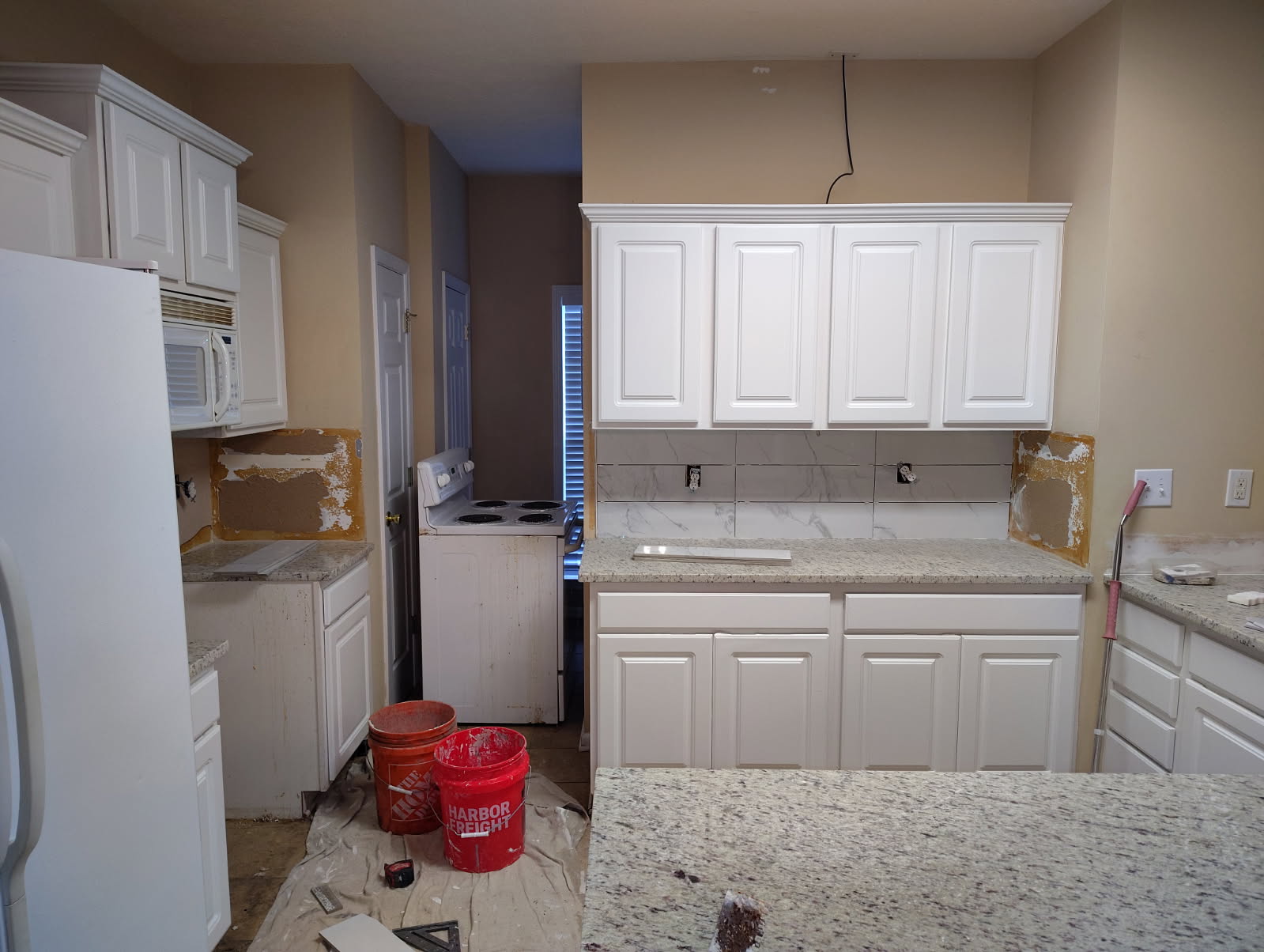 Kitchen mid renovation with new white cabinets, speckled granite counters and backsplash tiles partly installed, buckets on a drop cloth