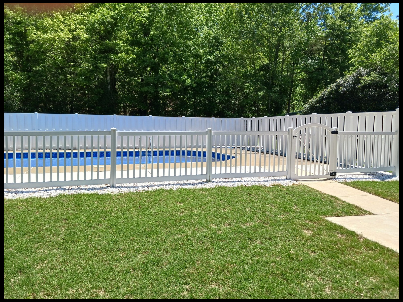 White vinyl privacy fence with vertical slats and an arched gate enclosing a swimming pool, with green trees behind