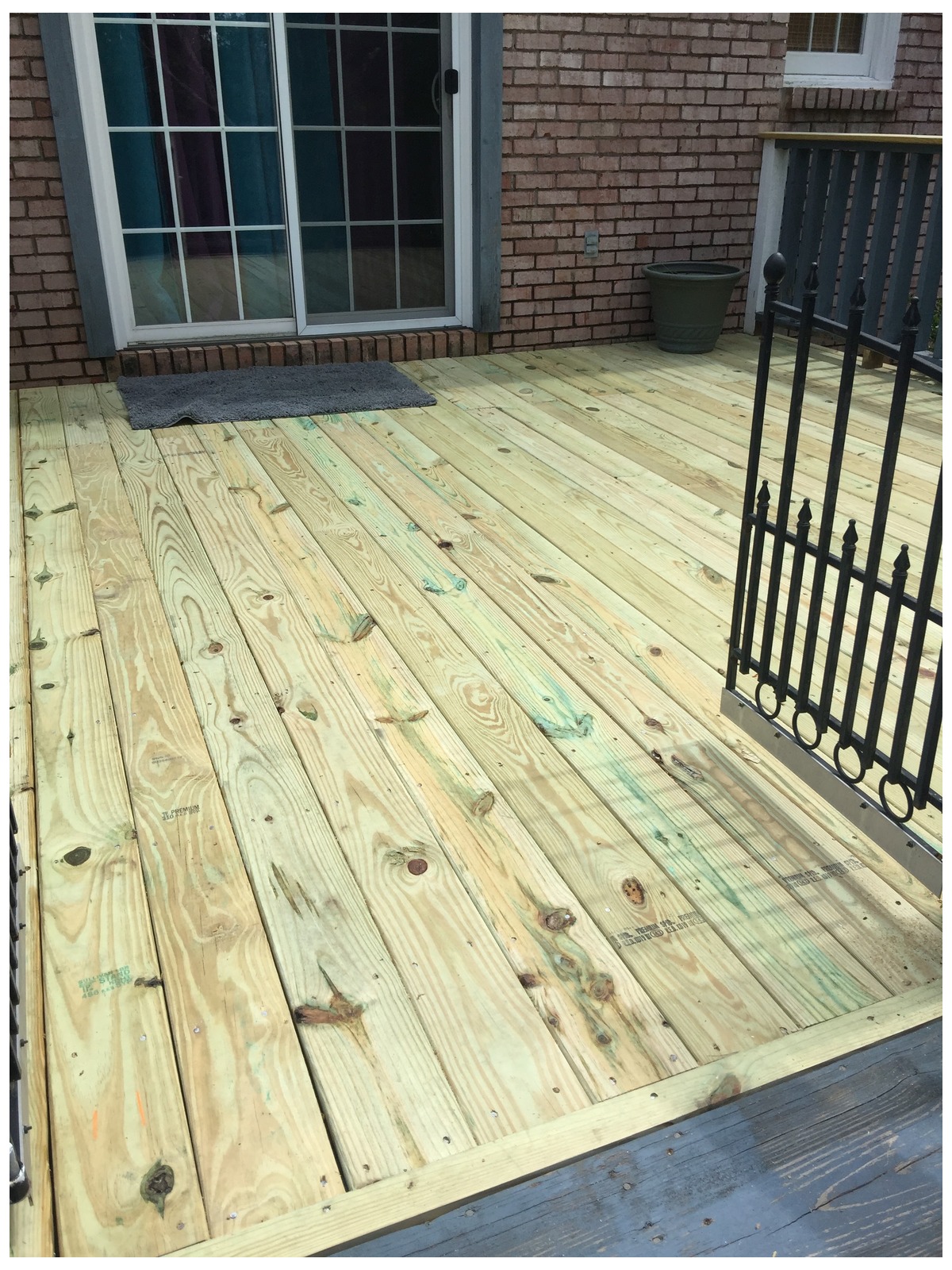 Newly laid pressure-treated deck boards against a brick wall with a white-framed glass door and a black metal railing