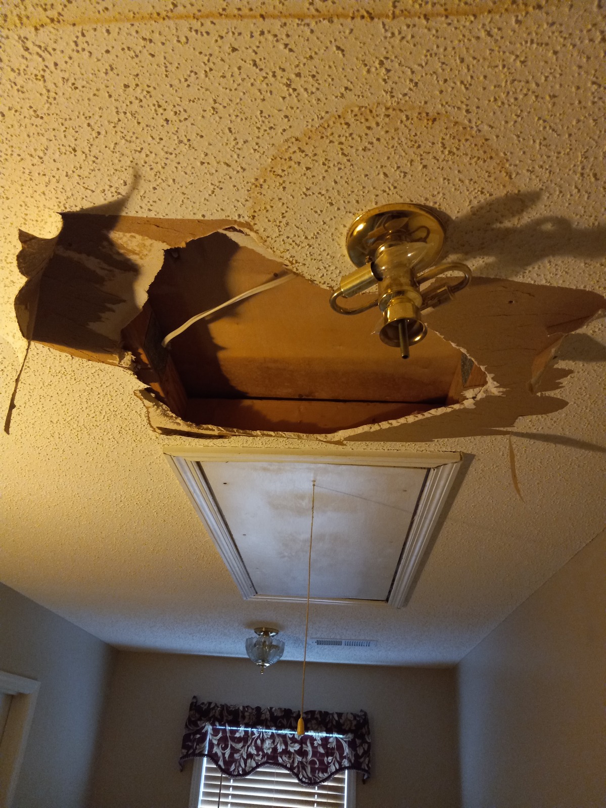 Damaged ceiling with a jagged hole in the drywall exposing a wooden beam and an electrical wire, beside a brass light fixture