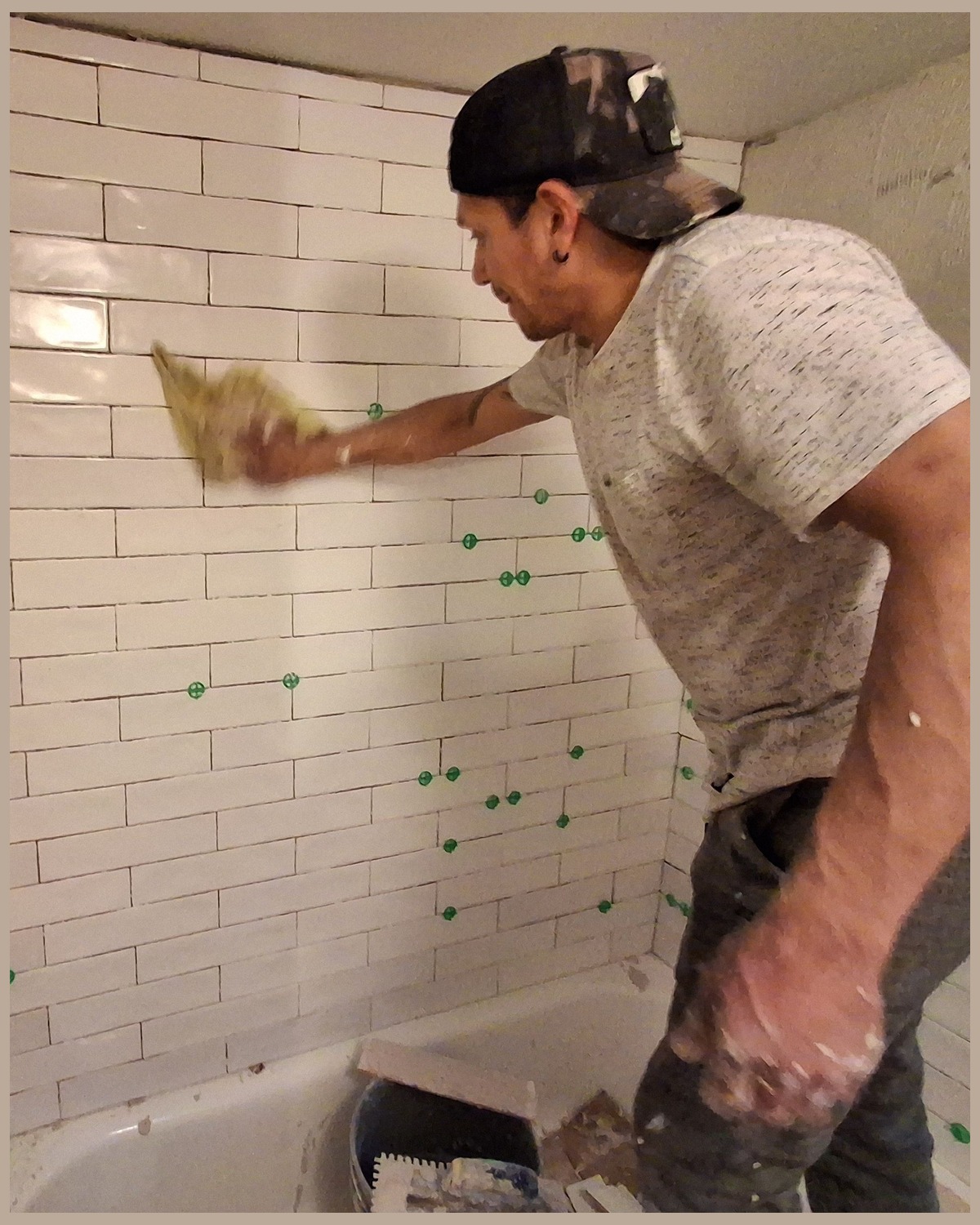 Handyman wiping newly set white wall tile with a yellow cloth in a bathroom, green spacers in the joints and a bathtub below
