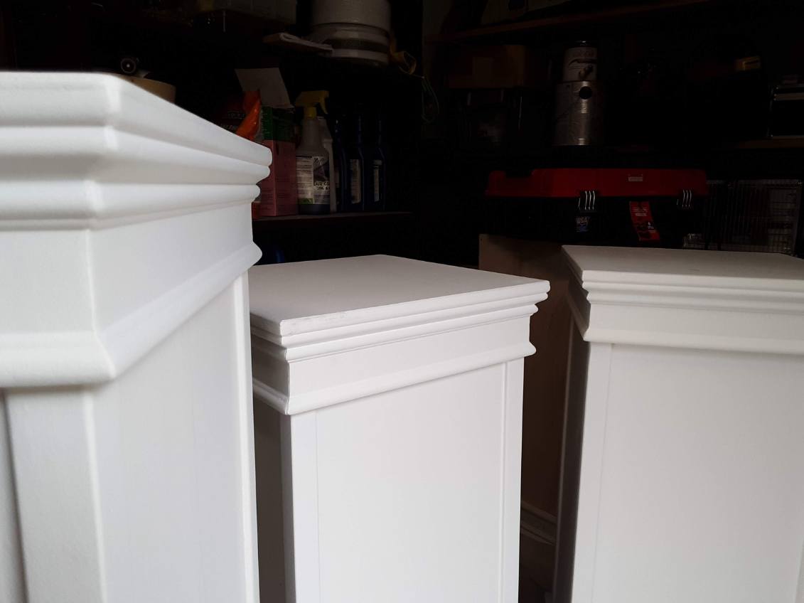 Three white painted column-style bases with molded tops standing in a workshop