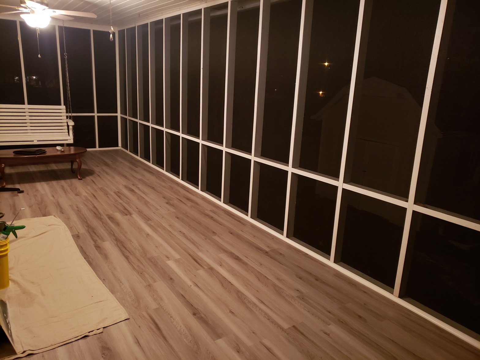 Screened-in porch at night with light wood-look plank flooring, white framing and dark screen panels, and a white hanging bench