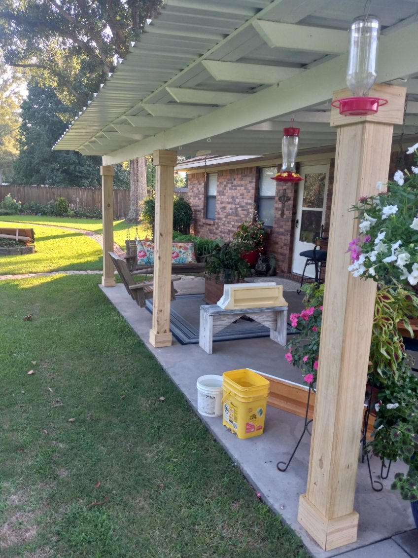 Backyard concrete patio under a corrugated metal roof on light wood posts, with a bench, a swing and potted plants