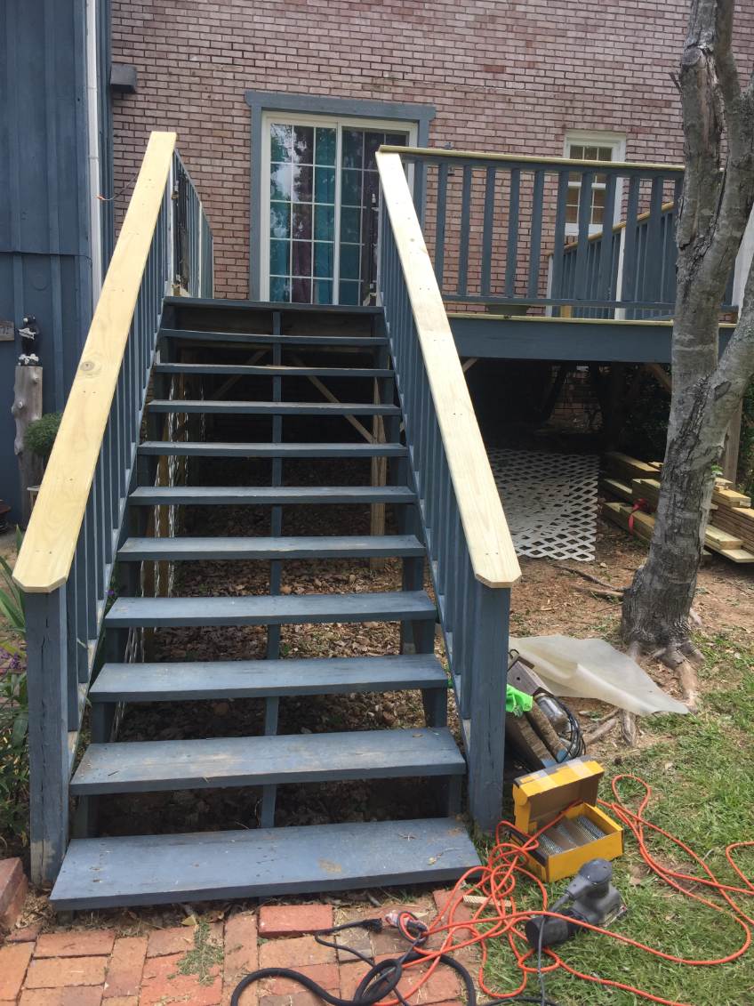 Newly finished dark gray wooden stairs with light wood handrails leading up to a deck on a brick building, tools on the ground below