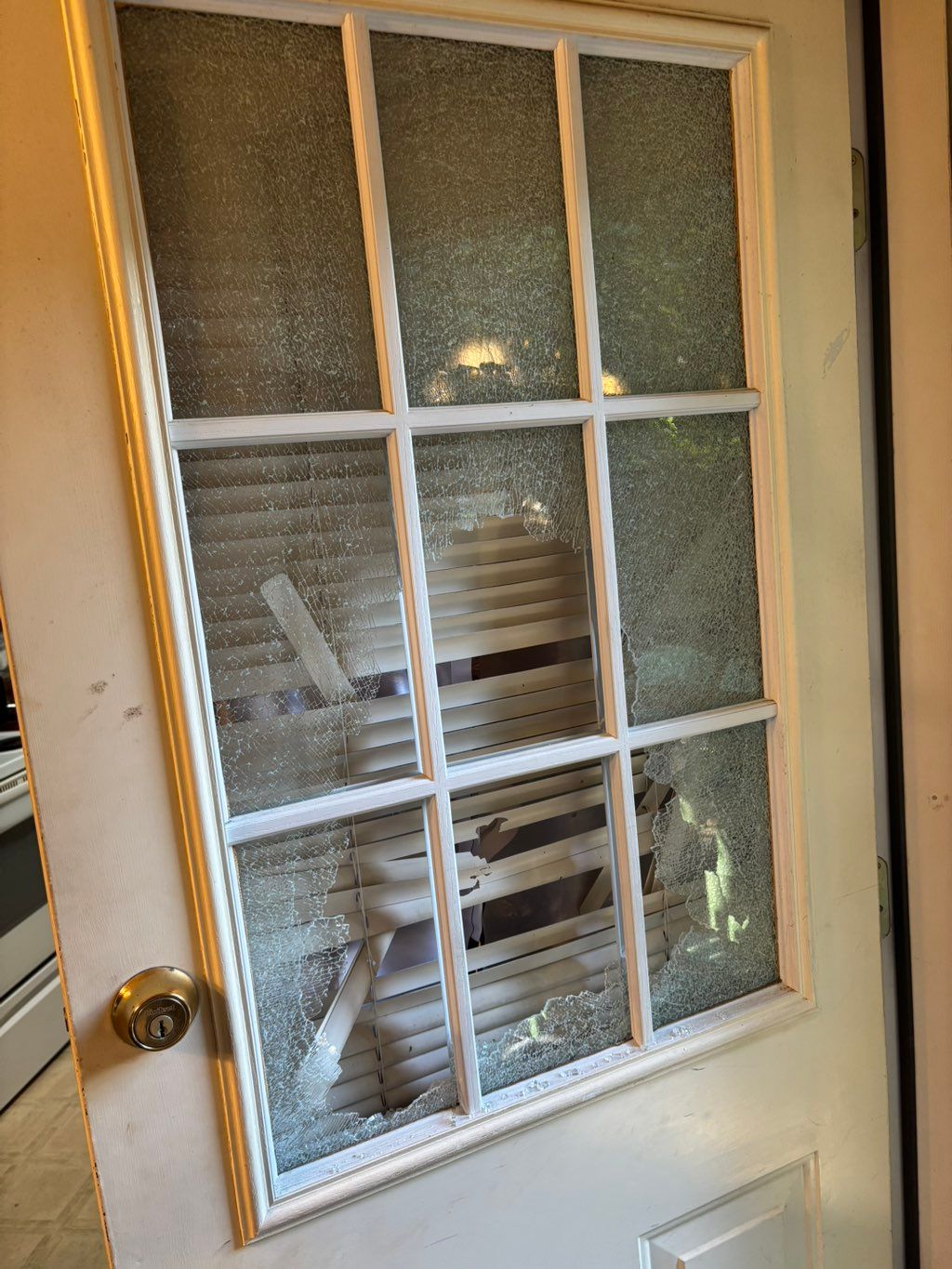 Shattered nine-pane glass in a white painted wooden door, exposing the blinds behind it